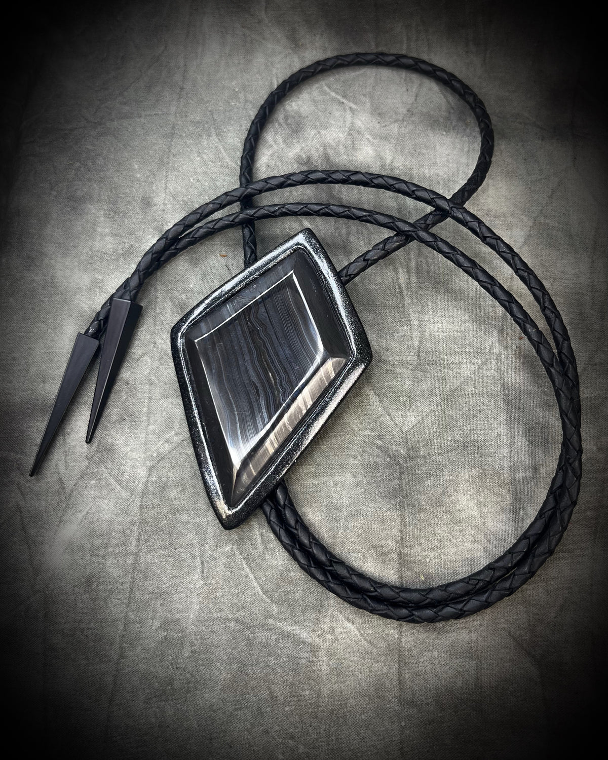 Genuine asymmetrical kite-shaped psilomelane bolo tie with black finish, black leather cord, and black spike bolo tips. Handmade by Become Spellbound.