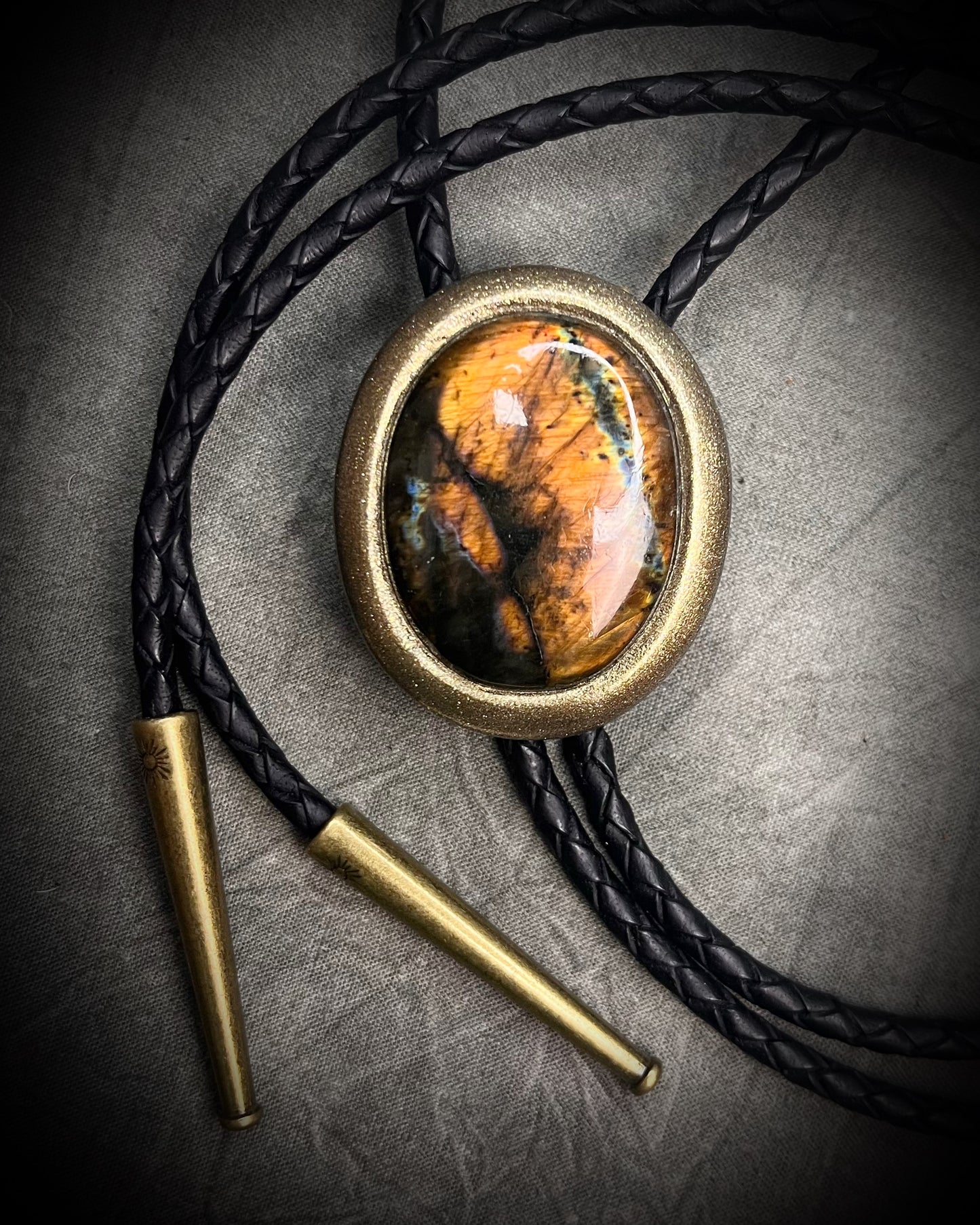 Glowing mystical orange labradorite bolo tie with blue stripe in a gold setting, black leather cord, and antique gold bolo tips. Handmade by Become Spellbound.