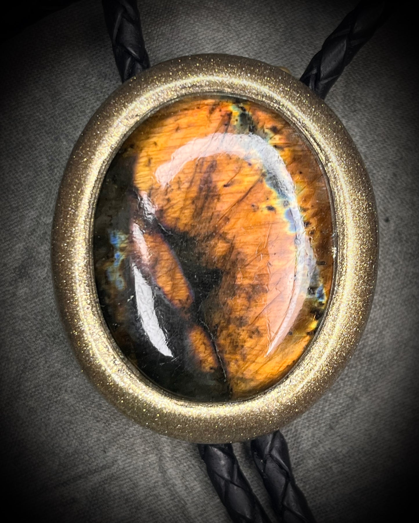 Glowing mystical orange labradorite bolo tie with blue stripe in a gold setting, black leather cord, and antique gold bolo tips. Handmade by Become Spellbound.