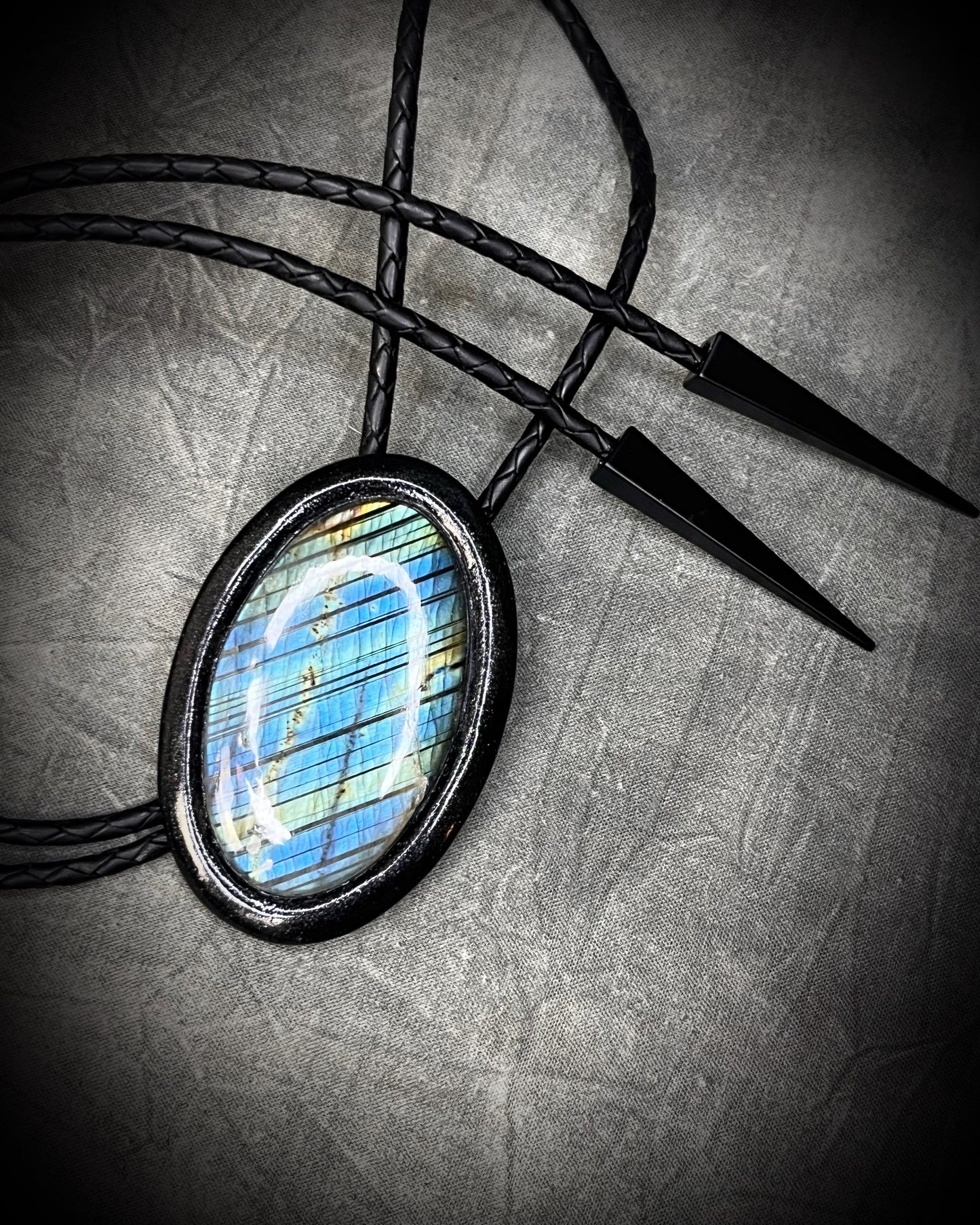 Handmade Bolo Tie Necklace with a blue and green labradorite pendant in black frame and leather cord on a textured gray background. 2026 Gothic western bolo trend in men's jewelry.