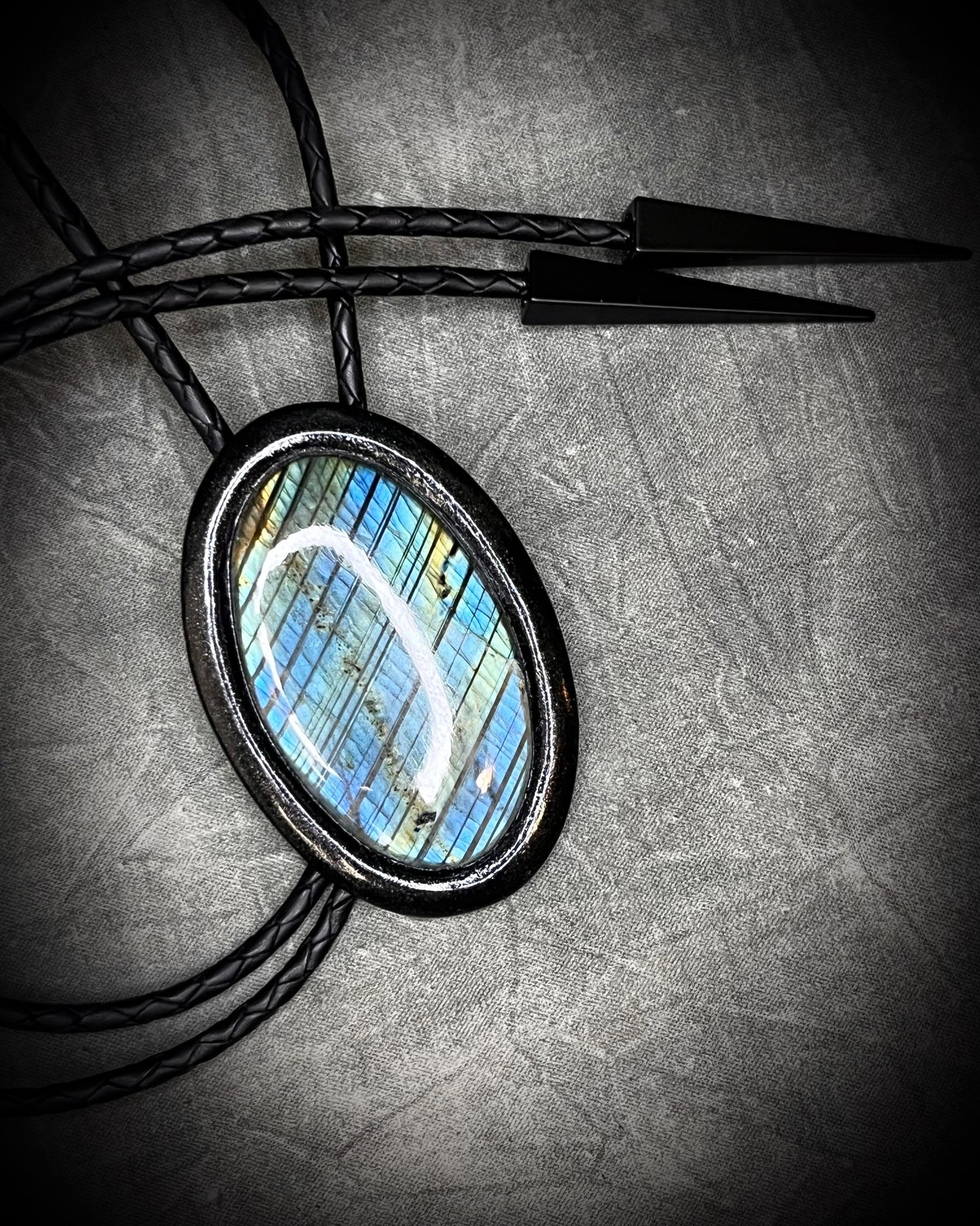 Handmade Bolo Tie Necklace with a blue and green labradorite pendant in black frame and leather cord on a textured gray background. 2026 Gothic western bolo trend in men's jewelry.
