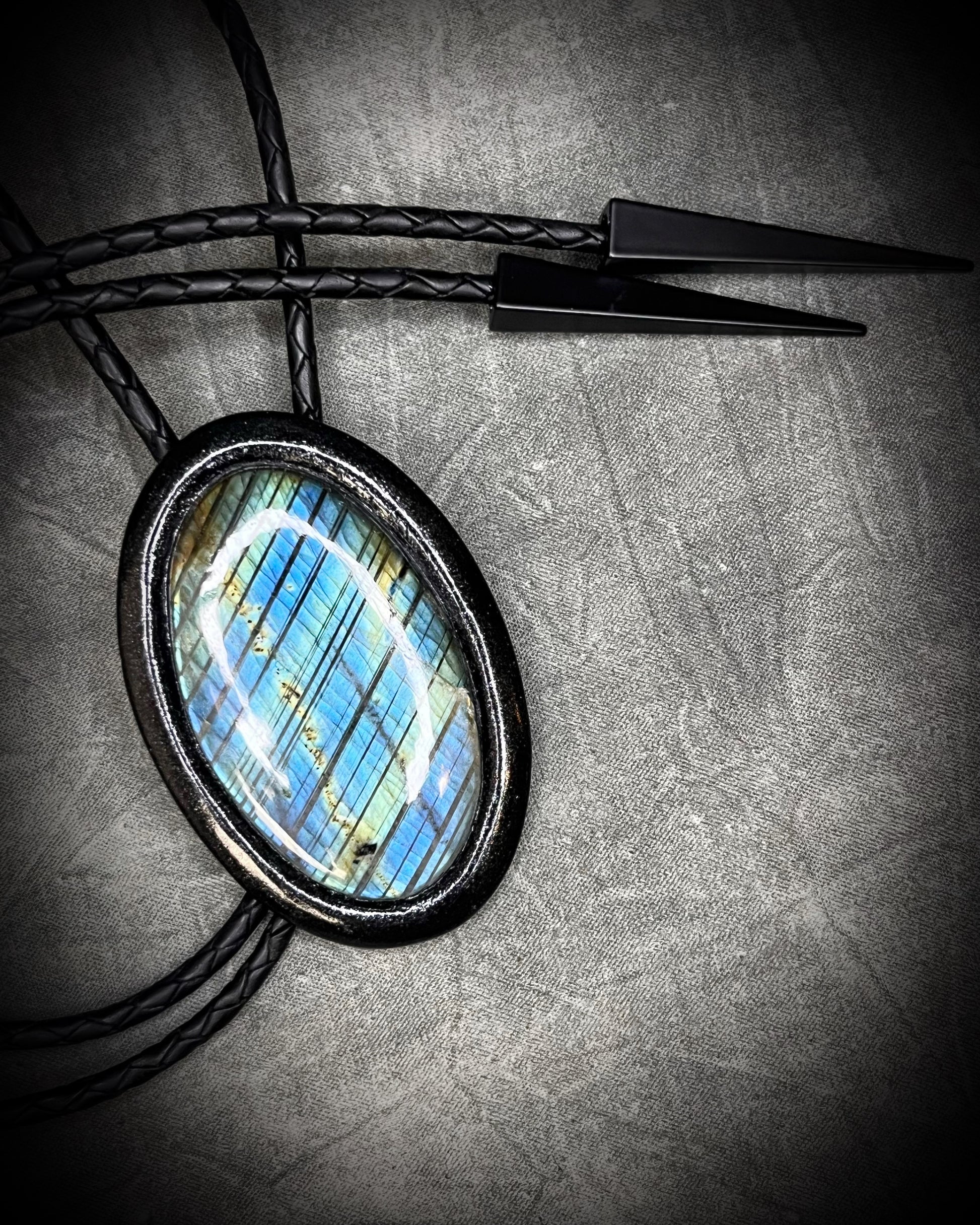 Handmade Bolo Tie Necklace with a blue and green labradorite pendant in black frame and leather cord on a textured gray background. 2026 Gothic western bolo trend in men's jewelry.