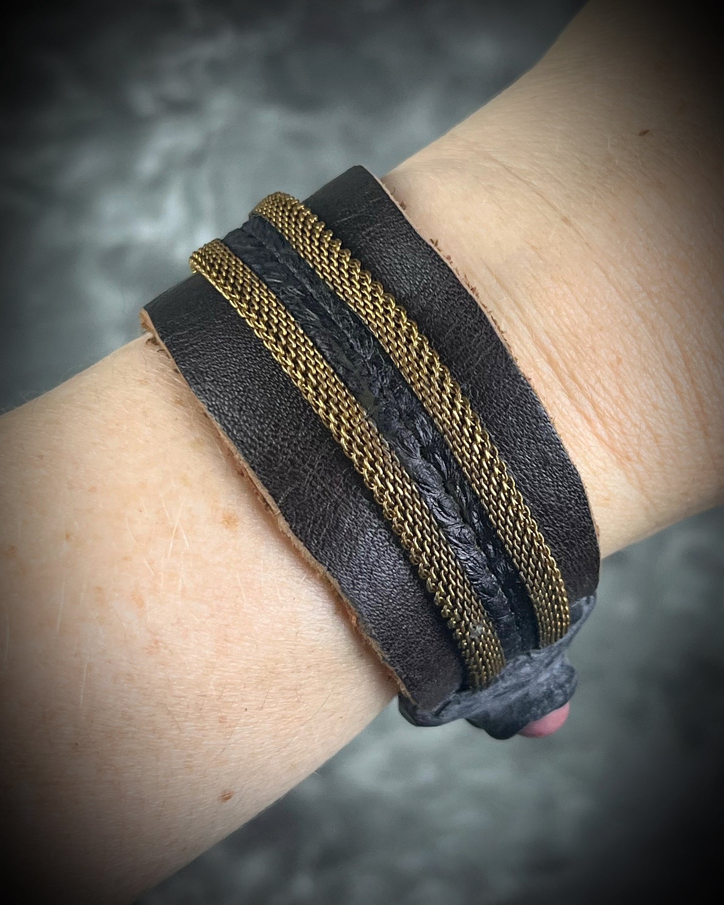 Handmade brown leather cuff bracelet with woven brass chain, rhodonite crystal details, and adjustable leather tie closure by Anothai Hansen.