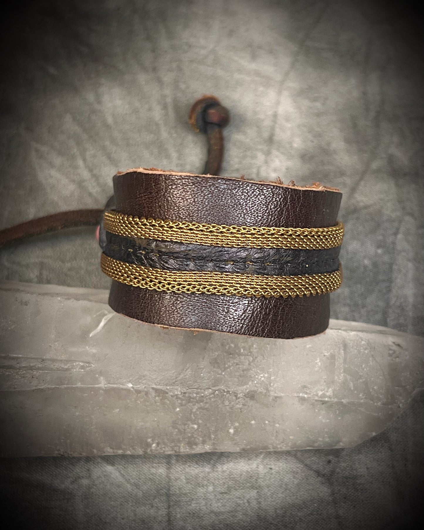 Handmade brown leather cuff bracelet with woven brass chain, rhodonite crystal details, and adjustable leather tie closure by Anothai Hansen.