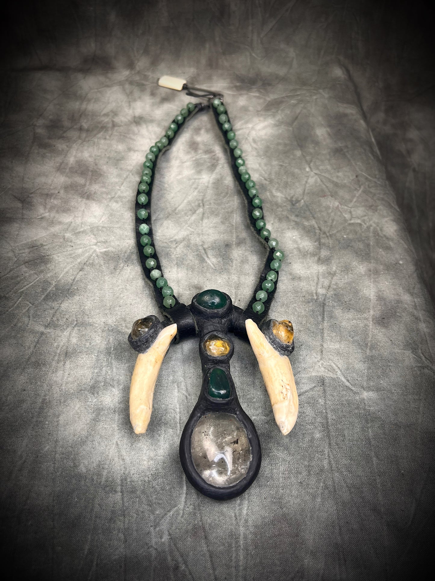 Handmade leather talisman with quartz, bloodstone, citrine, green aventurine, animal teeth, studded leather, and handmade wire clasp by Anothai Hansen.