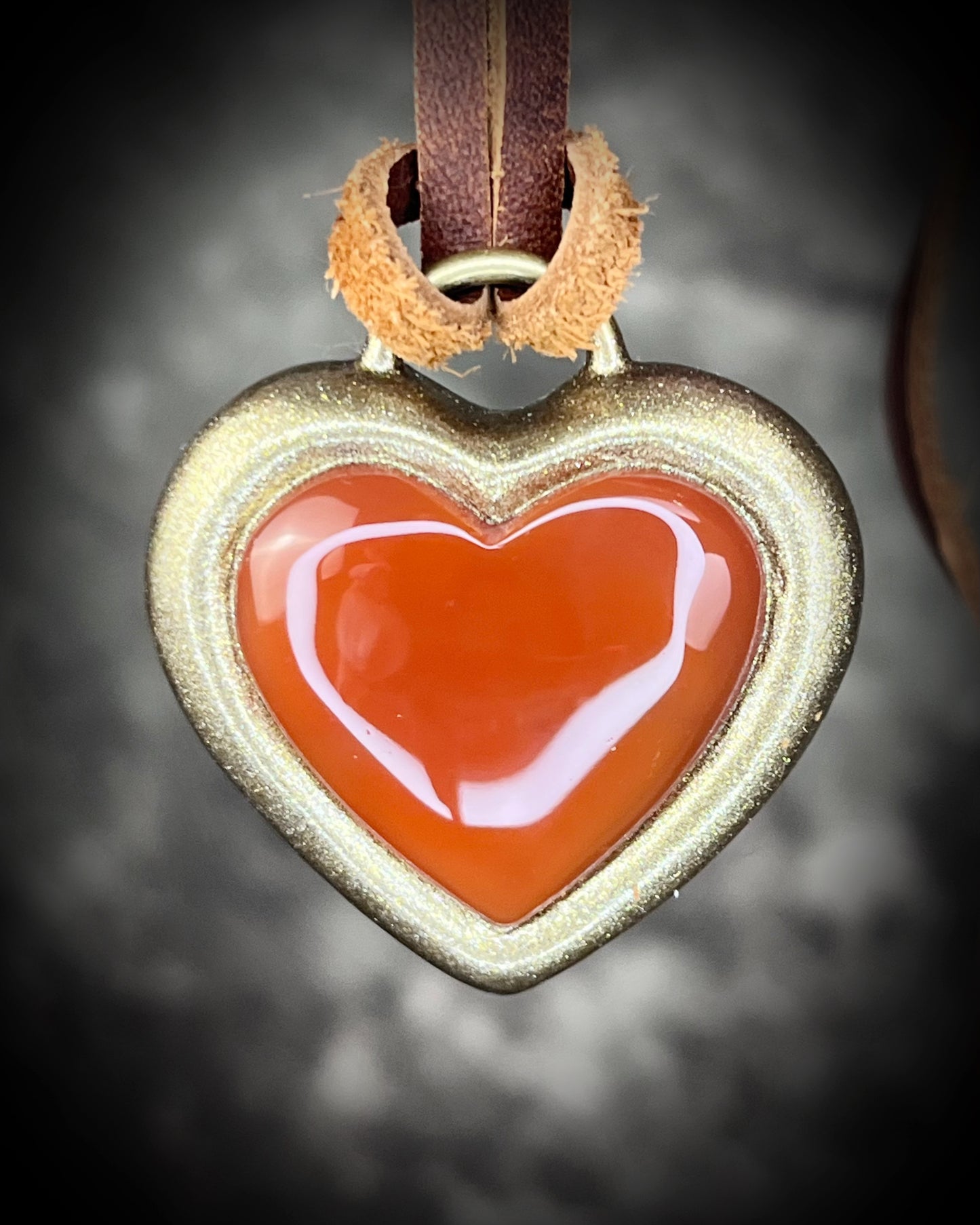 Heart-shaped pendant with a red center and gold frame, hung from brown leather on a dark background. Holiday gift for her!