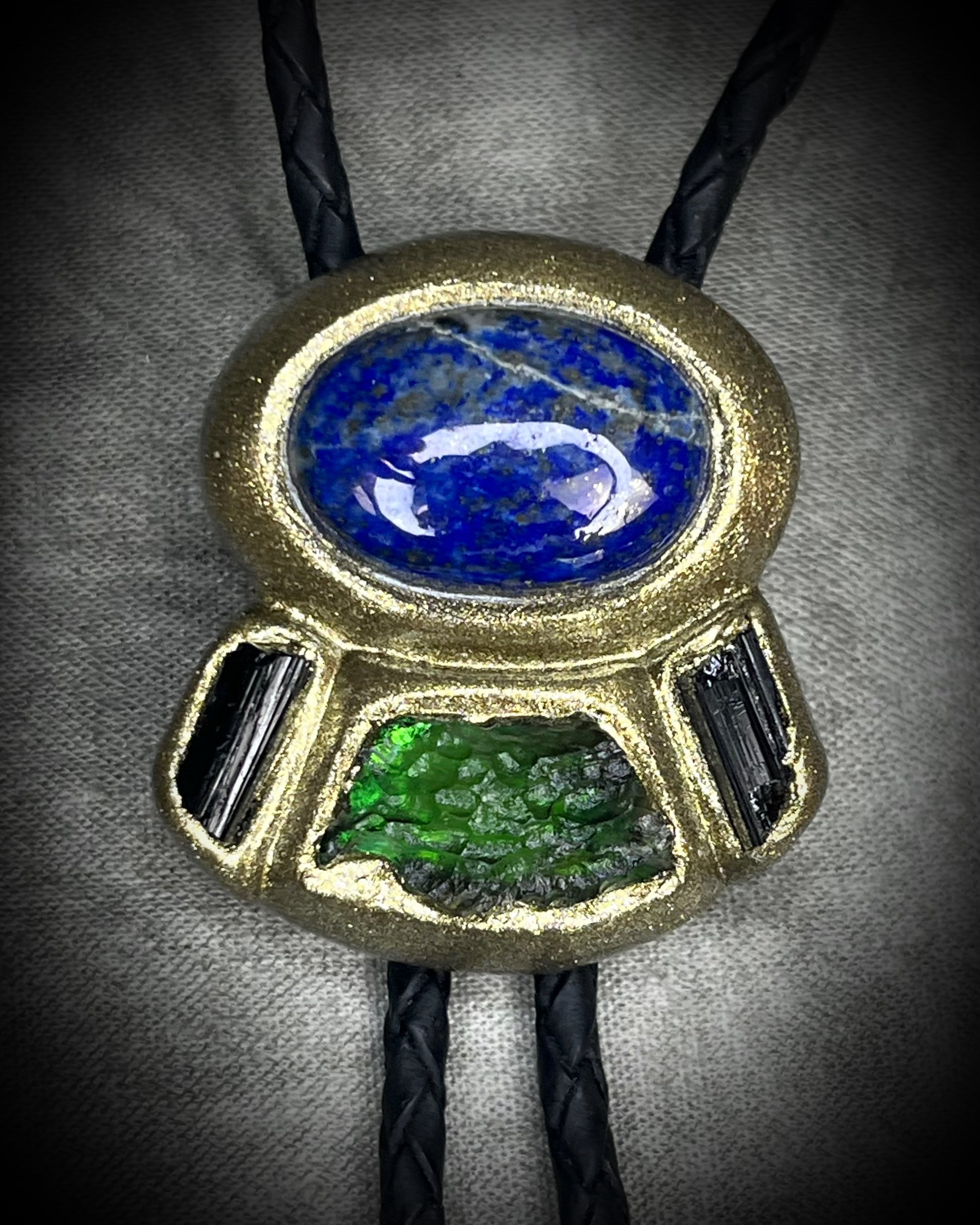 Moldavite bolo tie with lapis lazuli and black tourmaline on black leather, featuring gold finish and spike bolo tips, handmade by Become Spellbound.