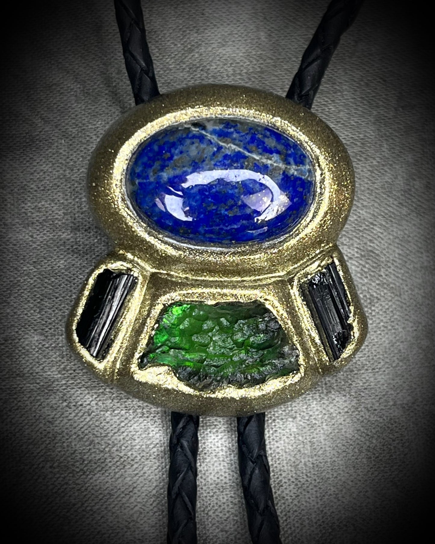 Moldavite bolo tie with lapis lazuli and black tourmaline on black leather, featuring gold finish and spike bolo tips, handmade by Become Spellbound.