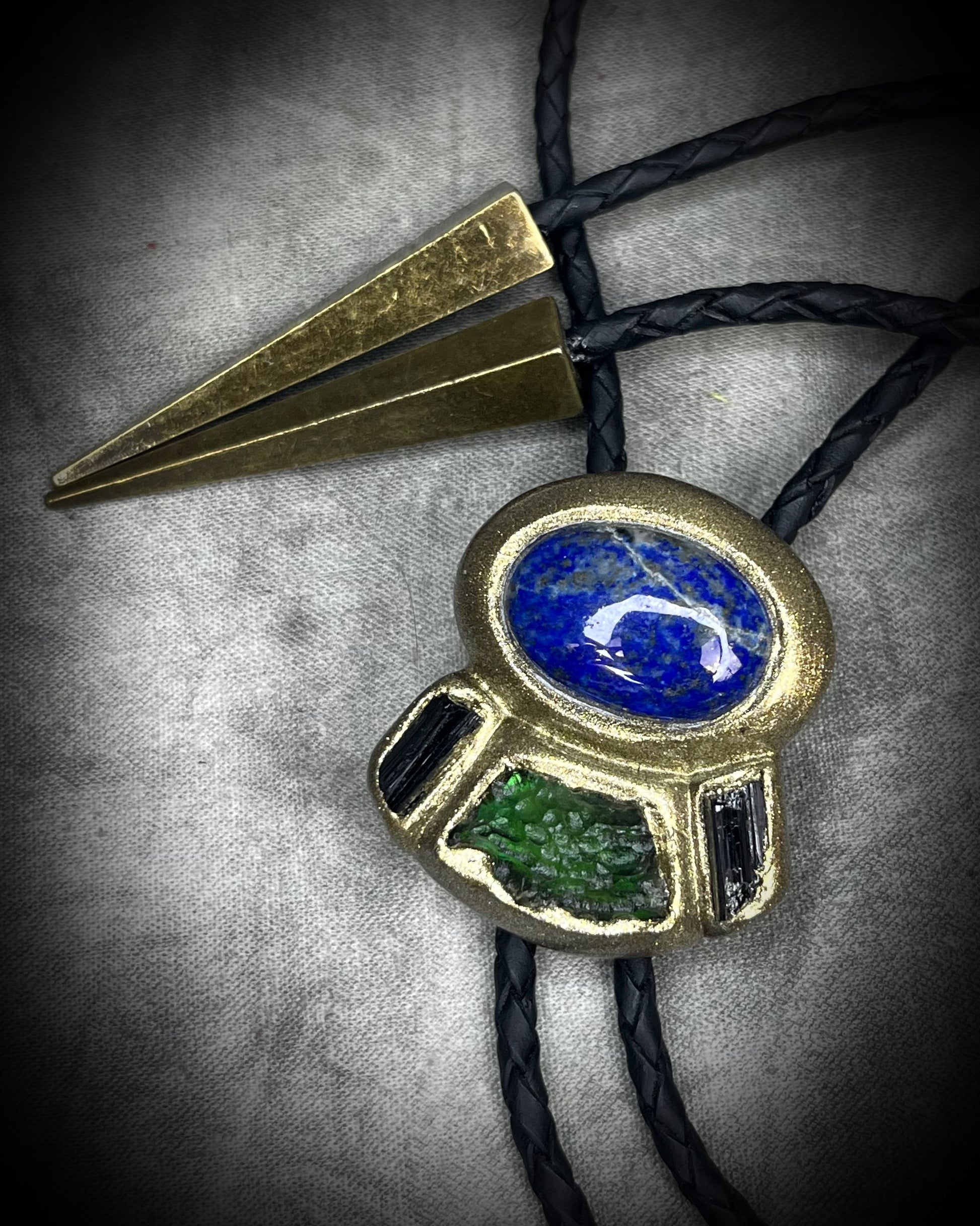 Moldavite bolo tie with lapis lazuli and black tourmaline on black leather, featuring gold finish and spike bolo tips, handmade by Become Spellbound.