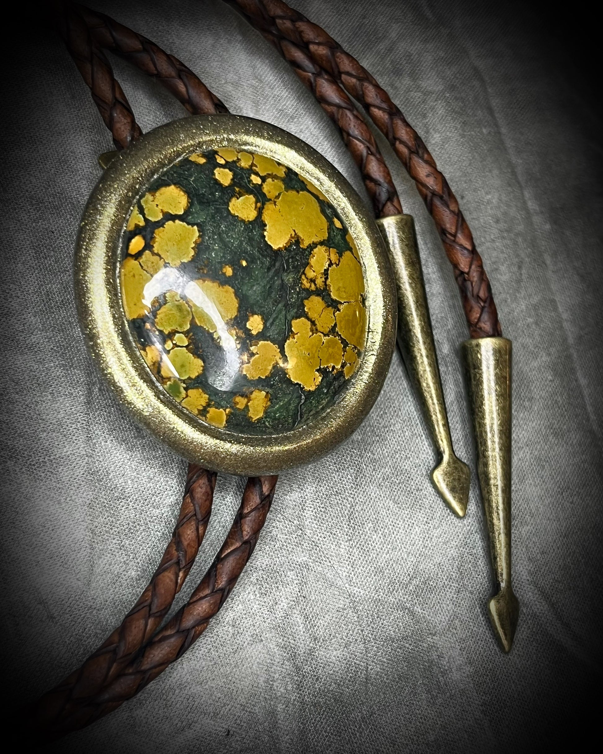 Mossy green and mustard-yellow turquoise bolo tie with natural plume markings, gold finish, brown leather, and gold spade tips.
