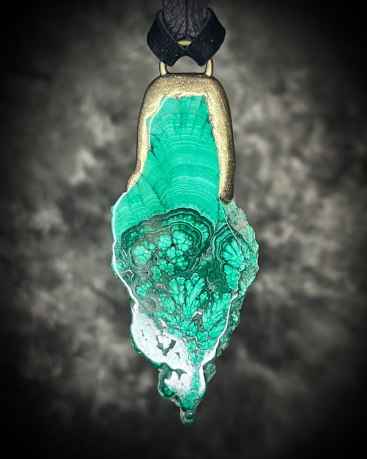 Natural malachite slab with distinctive green banding in a sculptural gold-finish talisman setting on black leather, handmade by Become Spellbound in Baltimore, Maryland.