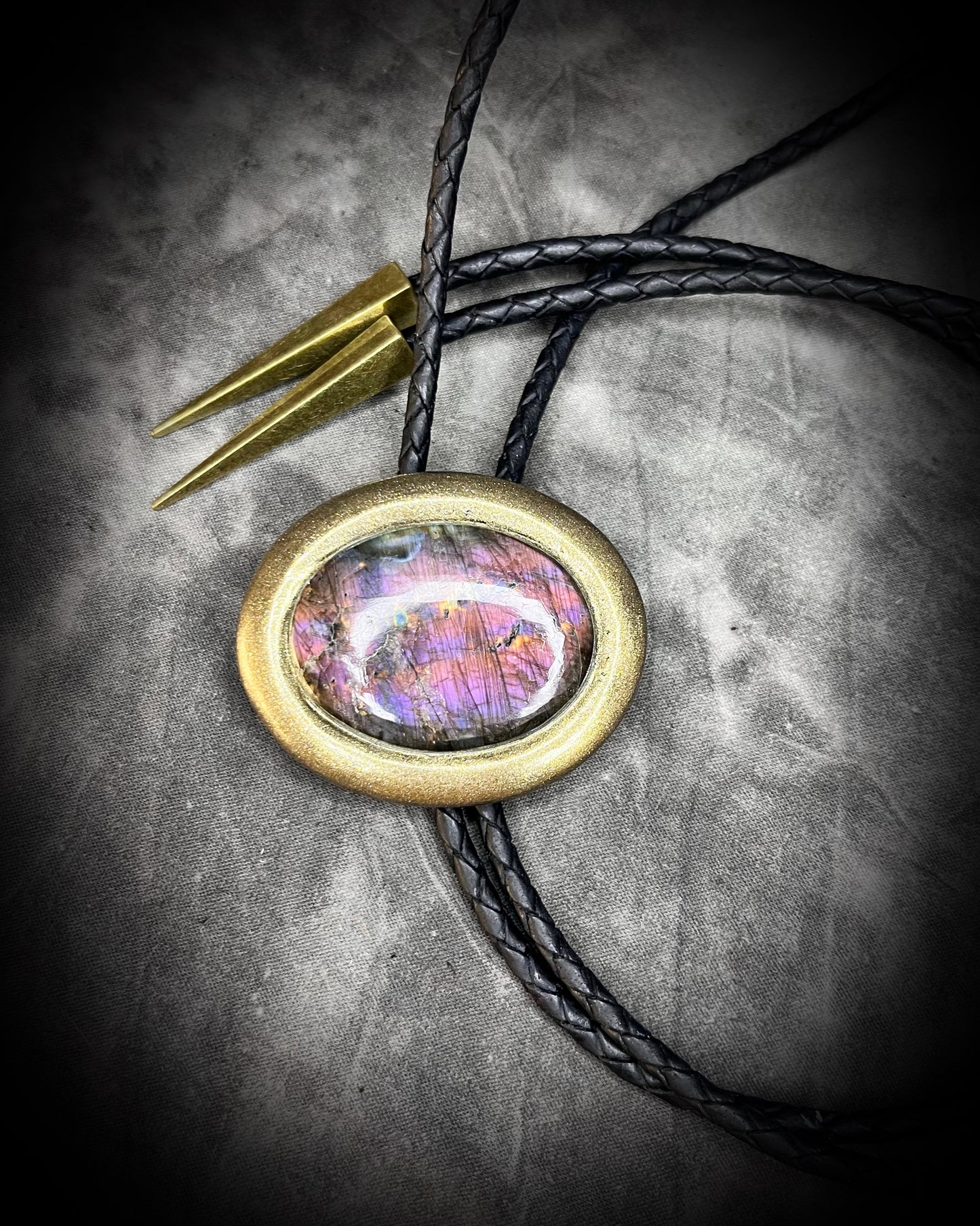 Necklace with a bolo tie design featuring a colorful purple labradorite stone pendant on a textured surface. Gold finish bolo tie with black leather, holiday gift for him.