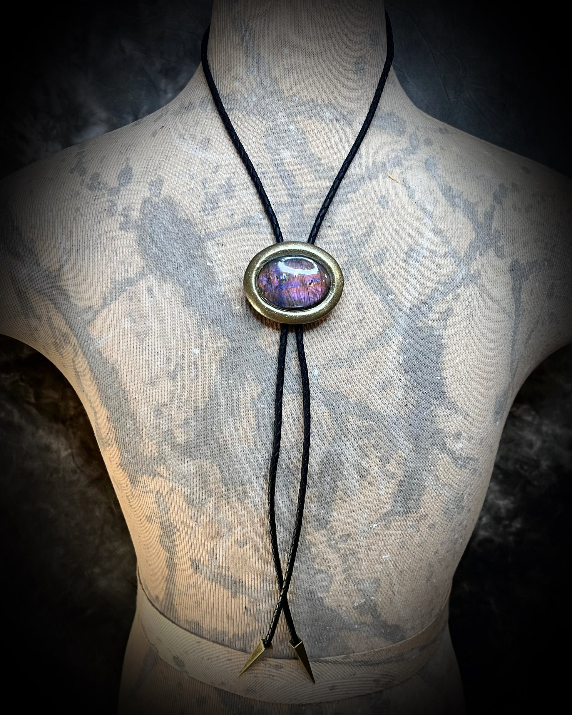 A male model bust wearing a bolo tie pendant featuring a colorful purple labradorite stone pendant. Gold finish bolo tie with black leather, holiday gift for him.