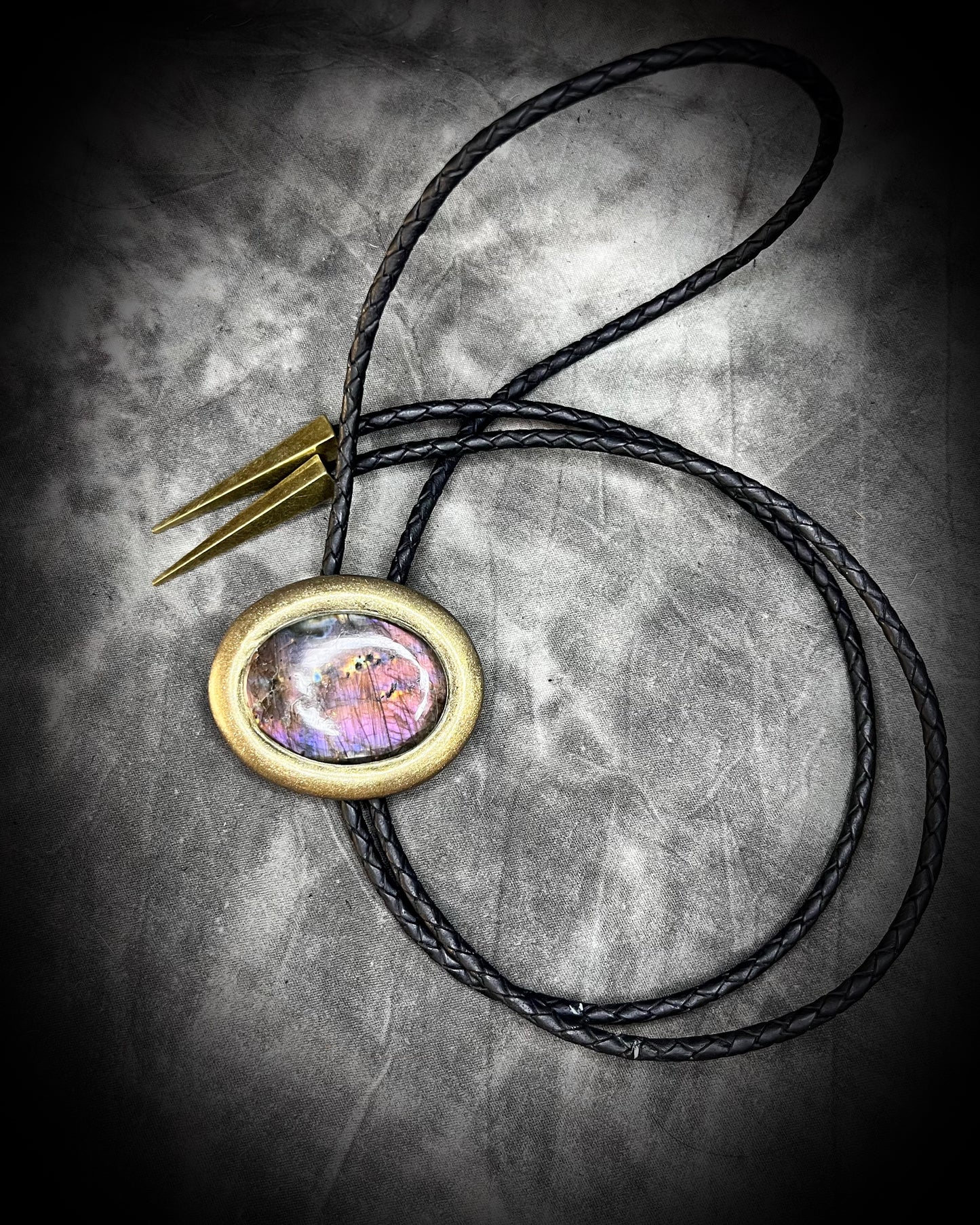 Necklace with a bolo tie design featuring a colorful purple labradorite stone pendant on a textured surface. Gold finish bolo tie with black leather, holiday gift for him.