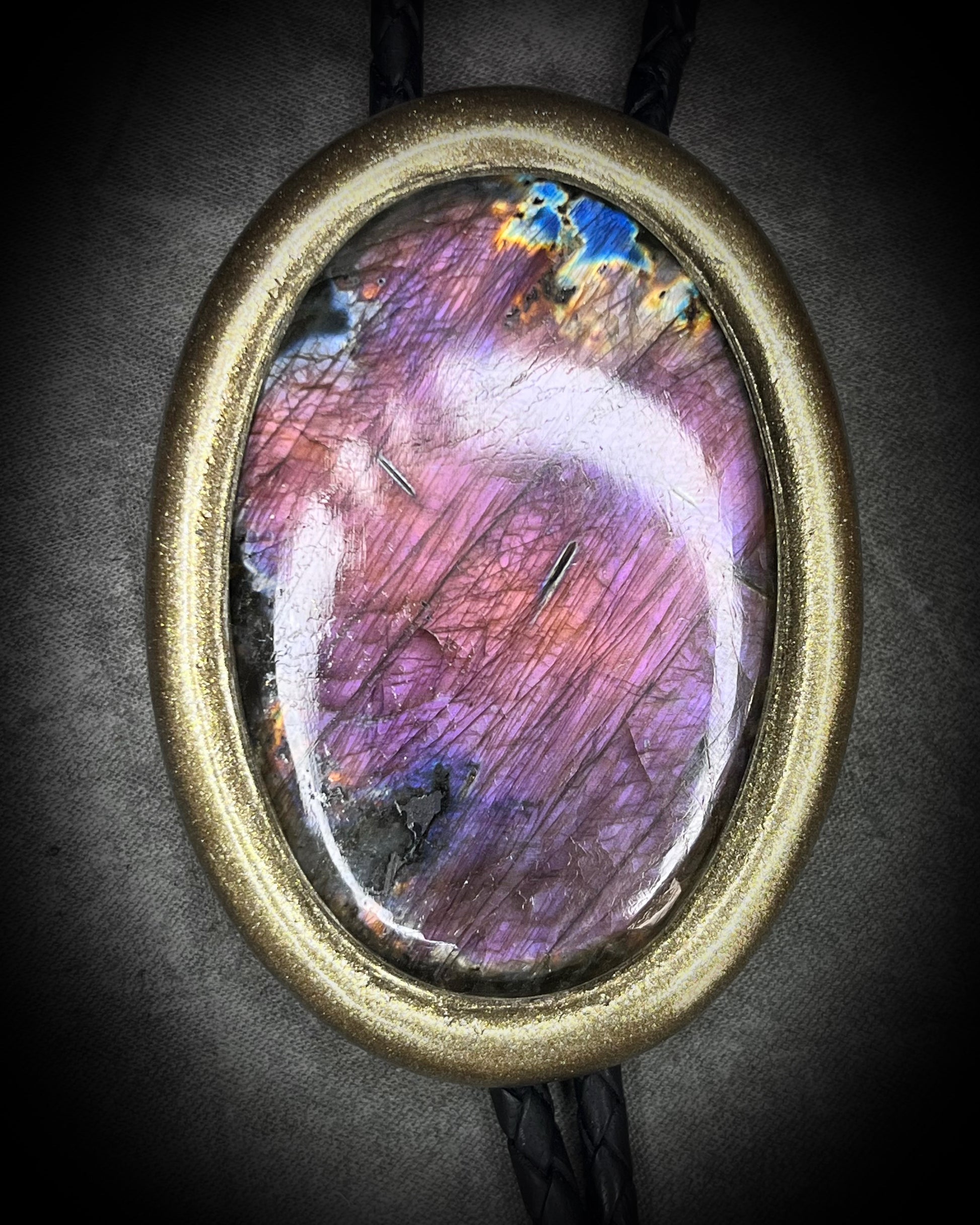 Oval-shaped purple labradorite gemstone pendant with purple and blue hues in a gold frame on a dark background. Handmade western bolo tie gift for him.