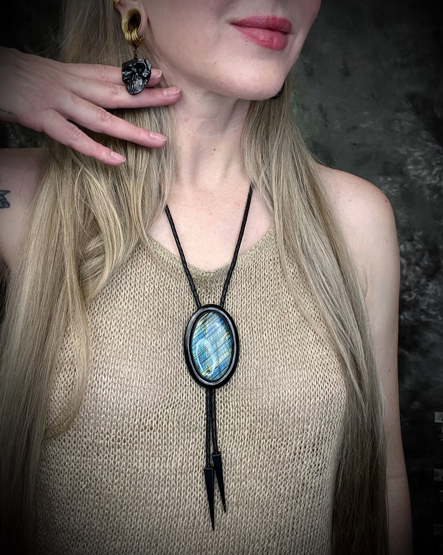 Person wearing a black framed bolo tie with a blue labradorite stone pendant against a dark background and skull shaped dangle earrings.