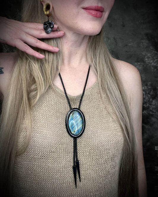Person wearing a black framed bolo tie with a blue labradorite stone pendant against a dark background and skull shaped dangle earrings.