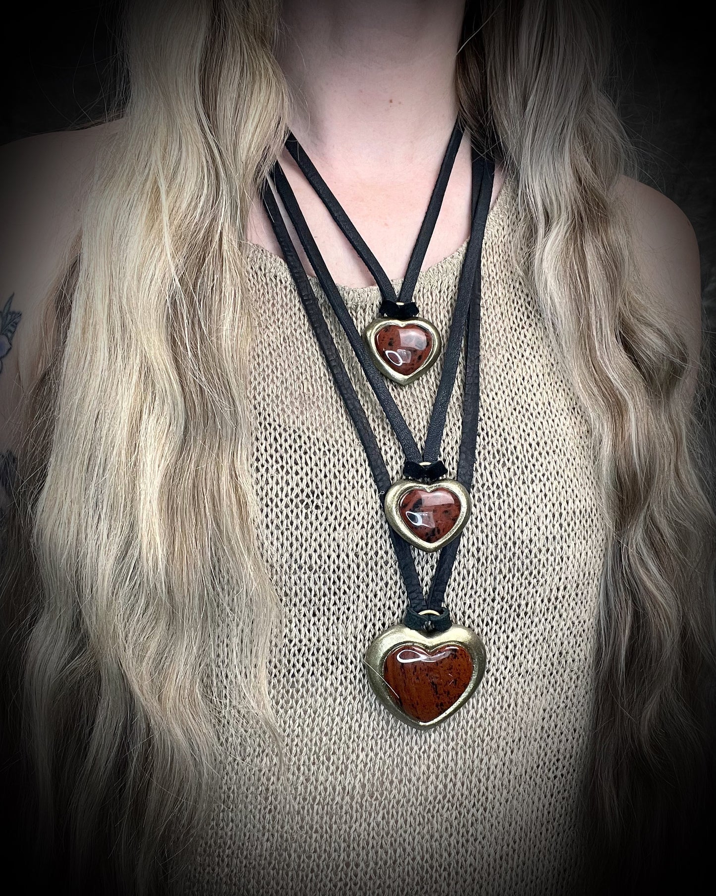 Person wearing multiple layering necklaces with heart-shaped red obsidian pendants on a dark background. Mahogany obsidian heart pendant necklace in gold finish on black leather.  Gift for her.