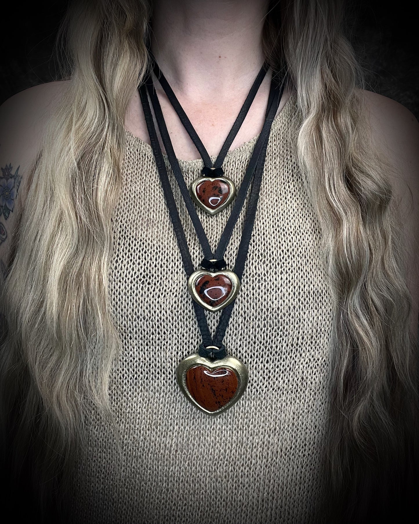 Person wearing multiple layering necklaces with heart-shaped red obsidian pendants on a dark background. Mahogany obsidian heart pendant necklace in gold finish on black leather.  Gift for her.