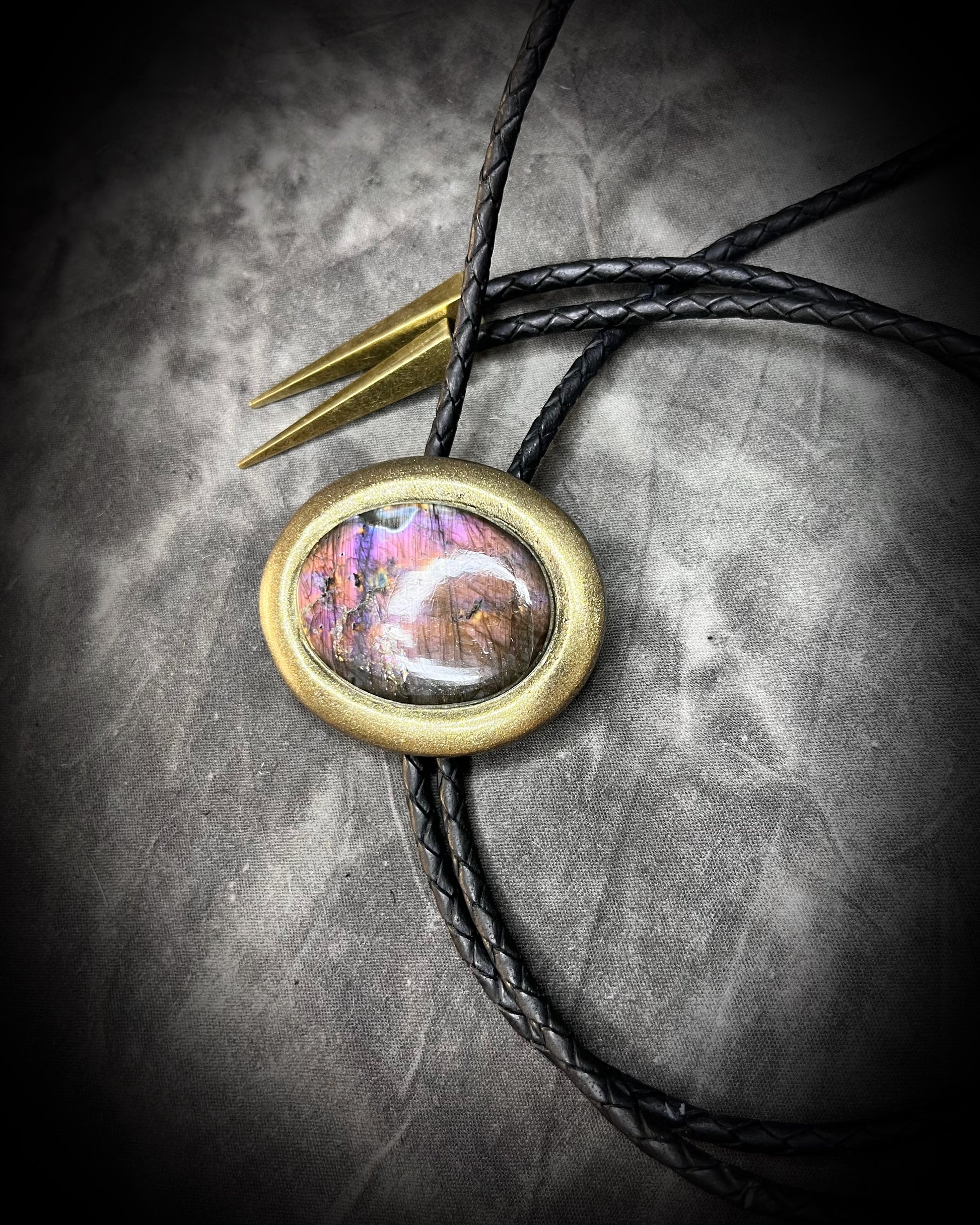 Purple labradorite Bolo tie with a colorful stone pendant and a gold finish on black leather on a textured surface.