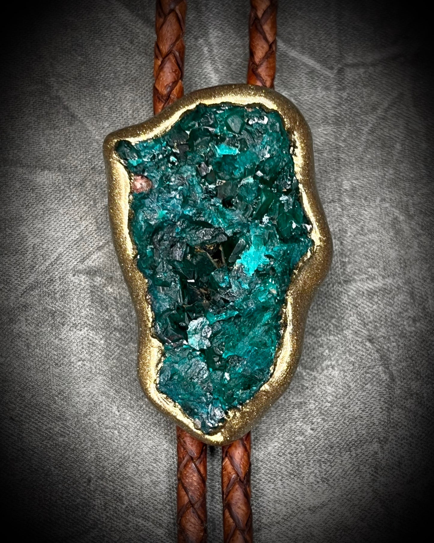 Closeup of Raw Dioptase teal colored gemstone Bolo tie set in a gold finish and mounted on earthy brown leather. A talisman for heart chakra healing, crystal jewelry for him. Western 2026 jewelry trend bolo. 