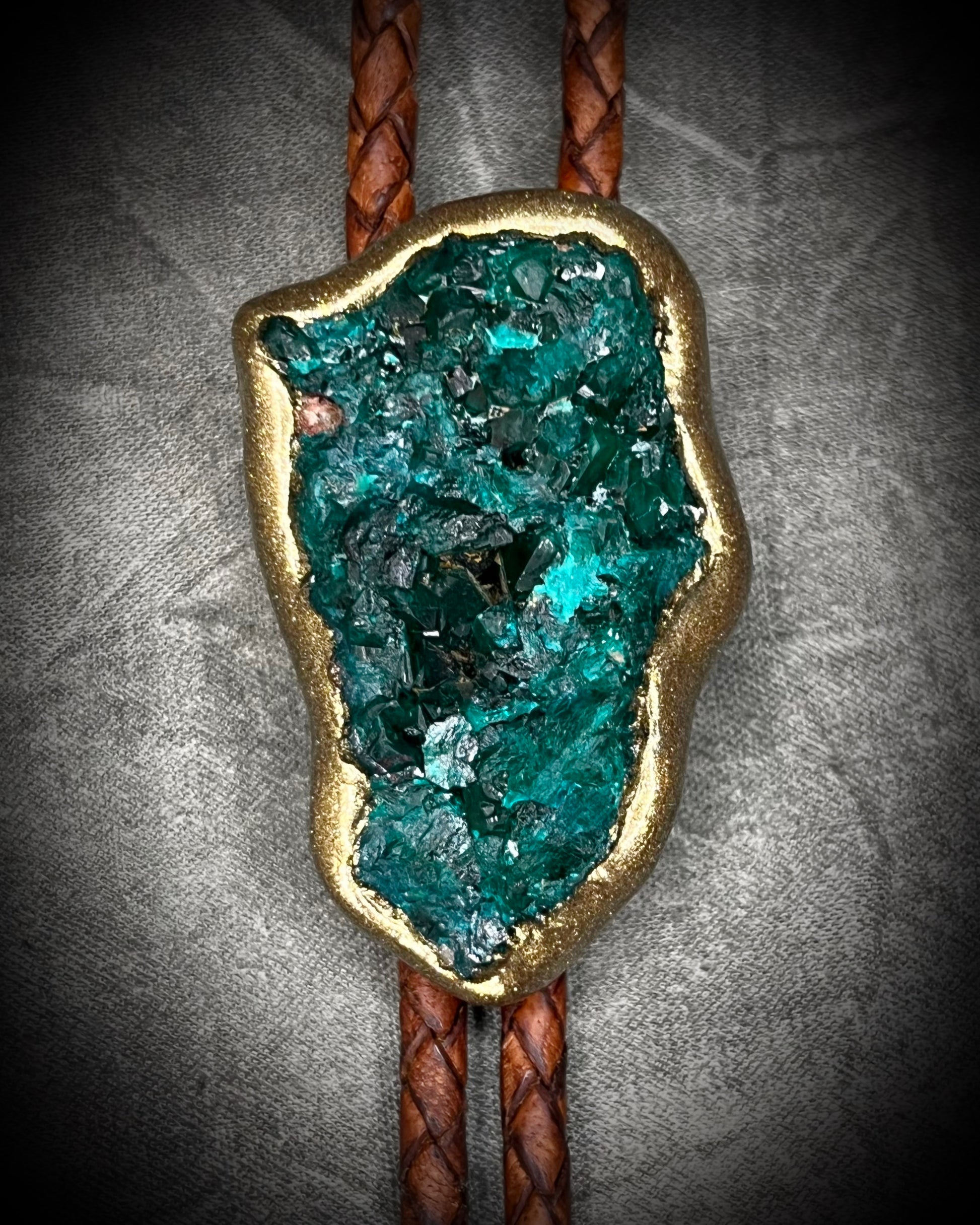 Closeup of Raw Dioptase teal colored gemstone Bolo tie set in a gold finish and mounted on earthy brown leather. A talisman for heart chakra healing, crystal jewelry for him. Western 2026 jewelry trend bolo. 