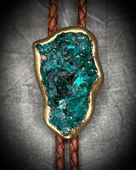 Closeup of Raw Dioptase teal colored gemstone Bolo tie set in a gold finish and mounted on earthy brown leather. A talisman for heart chakra healing, crystal jewelry for him. Western 2026 jewelry trend bolo. 