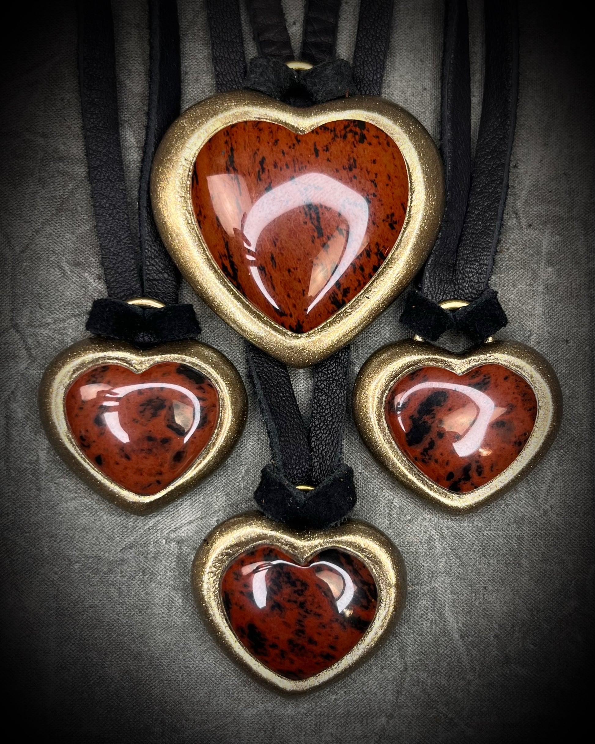 Several Mahogany obsidian heart pendant necklaces in gold finish on black leather in a flat lay arrangement of 4. Grounding protection crystal jewelry symbolizing strength and resilience. Gift for her.