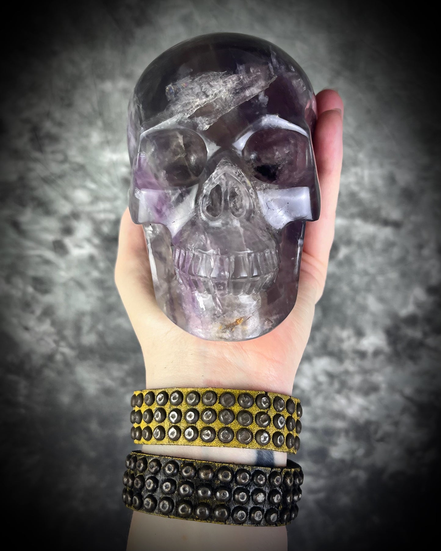 Two studded handmade leather bracelets worn layered on a wrist. Unisex rugged leather cuff jewelry with a crystal skull being held in the hand.