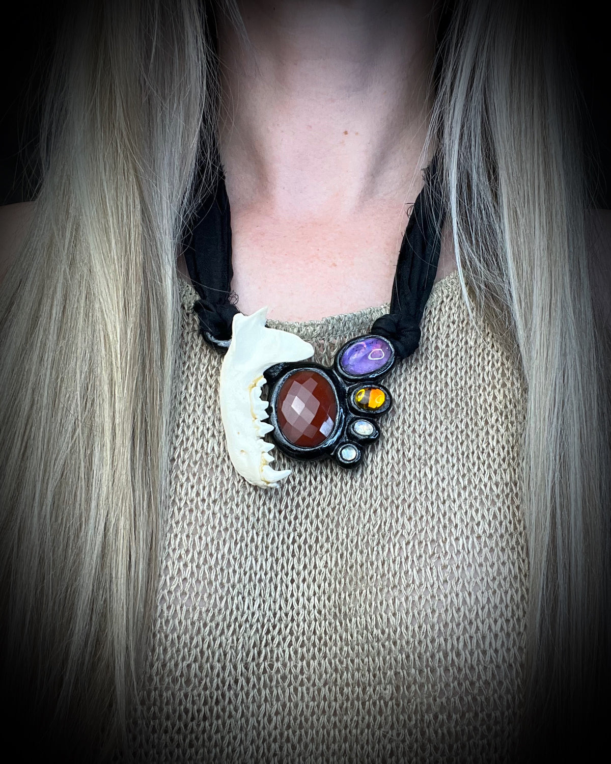 Otter jawbone talisman with a faceted carnelian center stone and amethyst, citrine, and moonstone accents in a sculptural black-finish setting on black recycled silk, handmade by Become Spellbound in Baltimore, Maryland.
