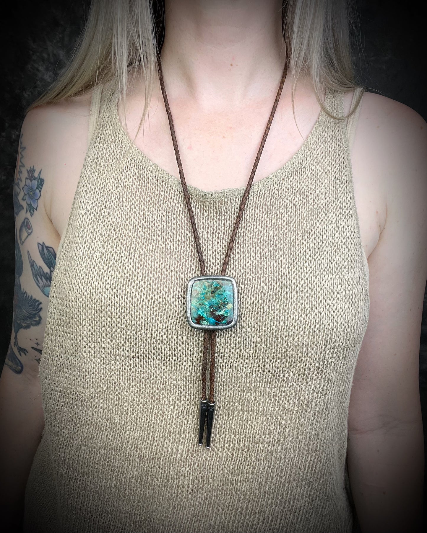 A person wearing a bolo-style necklace with a square turquoise-like pendant and dark brown cord, shown against a dark background.