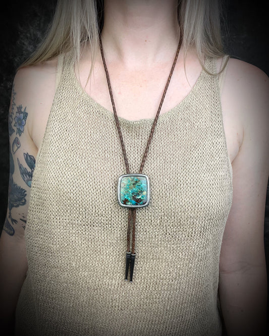 A person wearing a bolo-style necklace with a square turquoise-like pendant and dark brown cord, shown against a dark background.