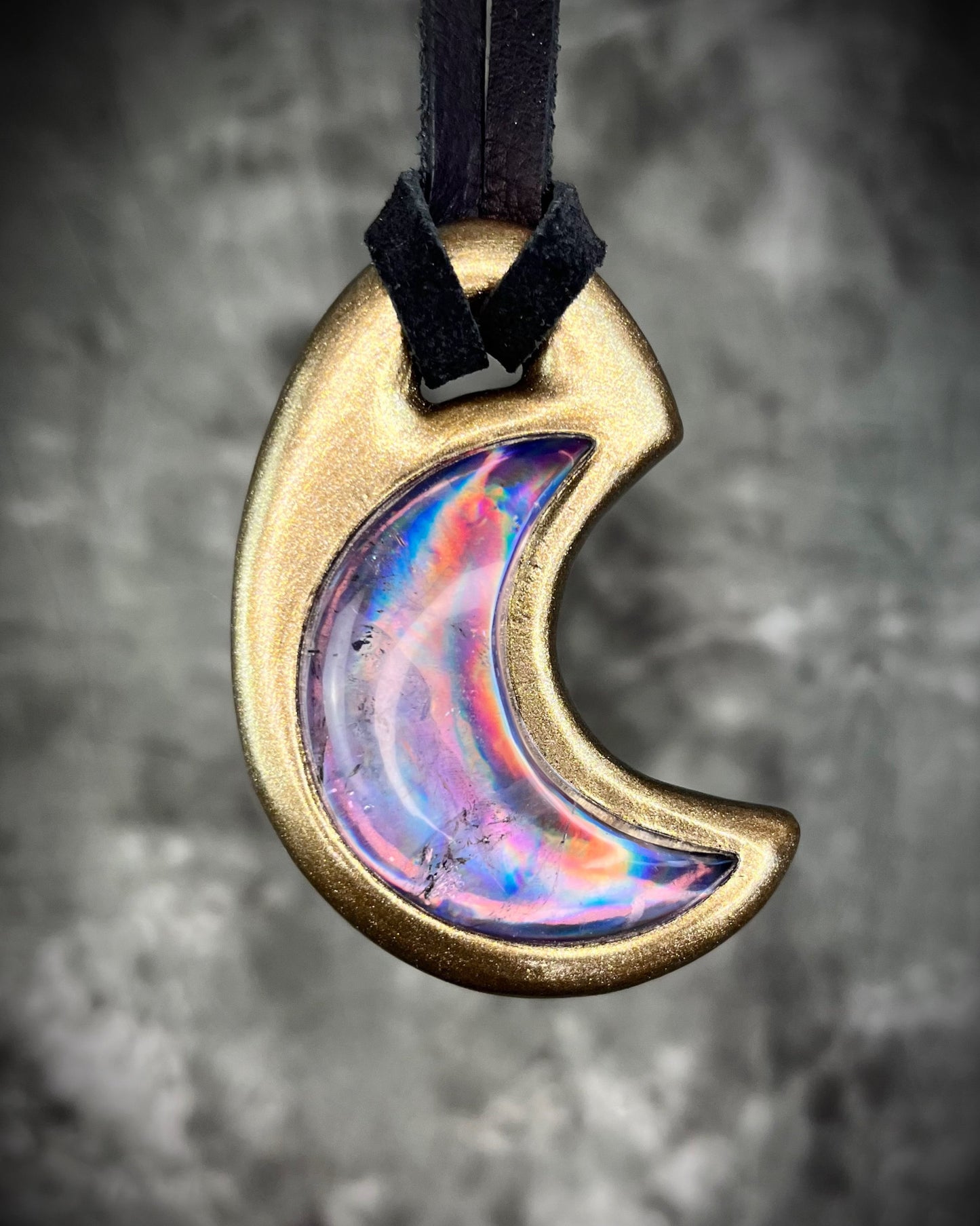 A crescent moon-shaped Bowie Black Rainbow Quartz Crescent Moon necklace with a leather strap, displaying a multicolored iridescent pattern.