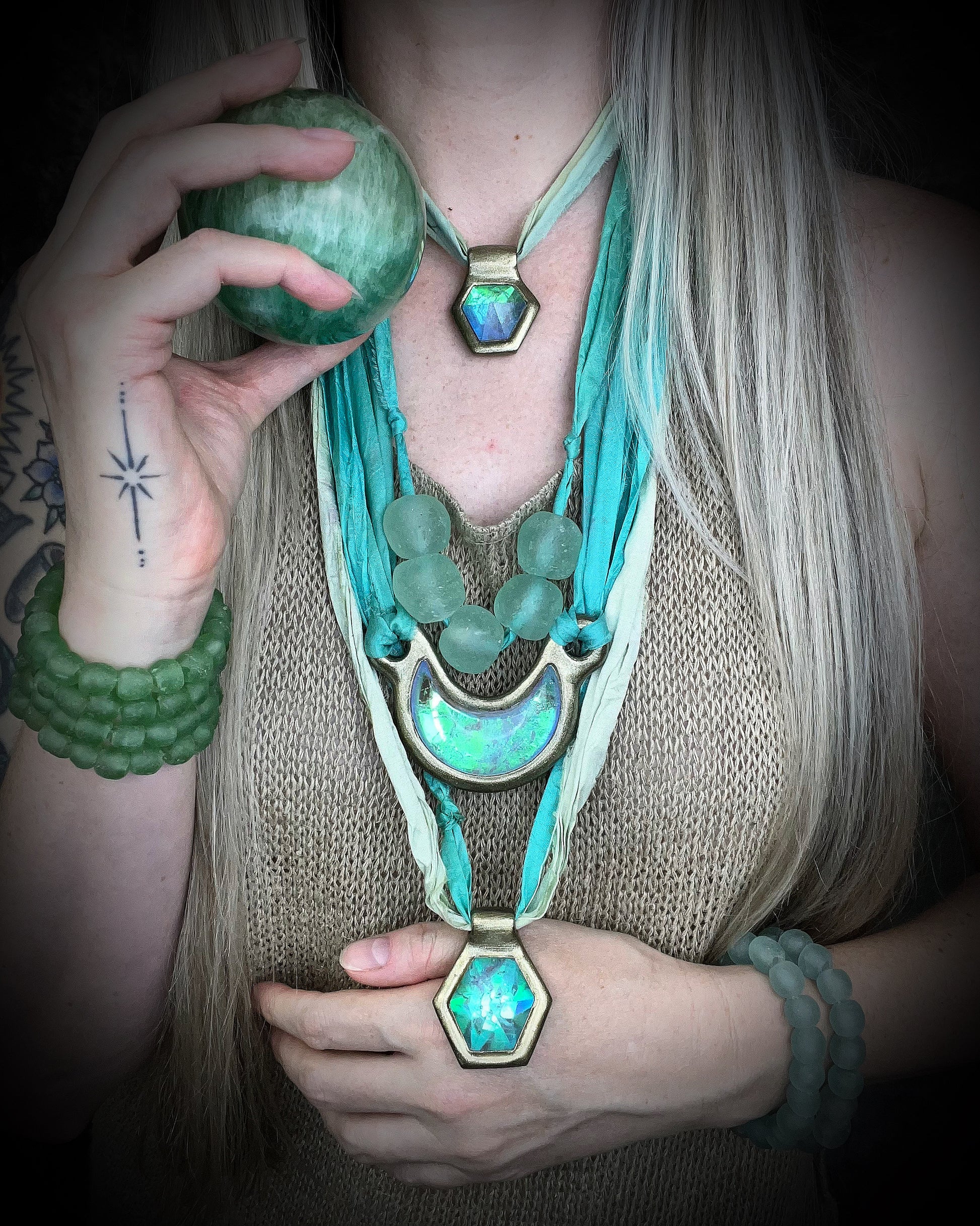 Model holding a green crystal ball and wearing recycled glass beaded necklace and bracelets, layered with quartz, chrysoprase and moon pendants with recycled silk in mint and aqua hues for a festival, summer or travel jewelry vibe!